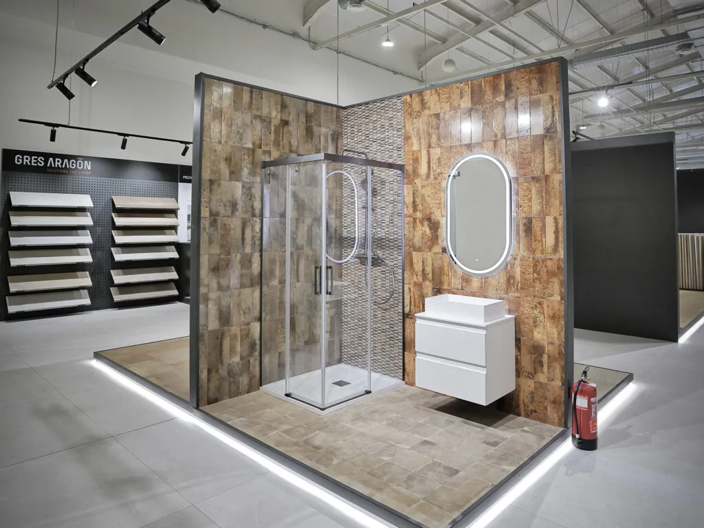 Ambiente de baño expuesto con mampara, revestimiento cobrizo y espejo ovalado en el showroom